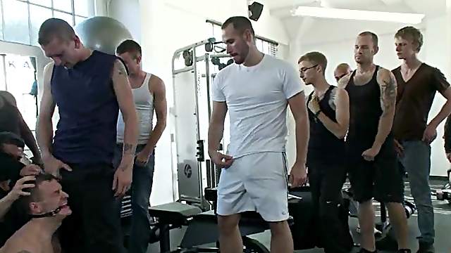 A tied up guy sucks huge dicks and get toyed in a gym